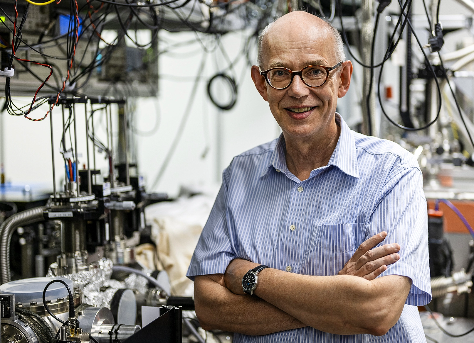 A man standing in front of an optical setup with his arms crossed smiles at the camera.