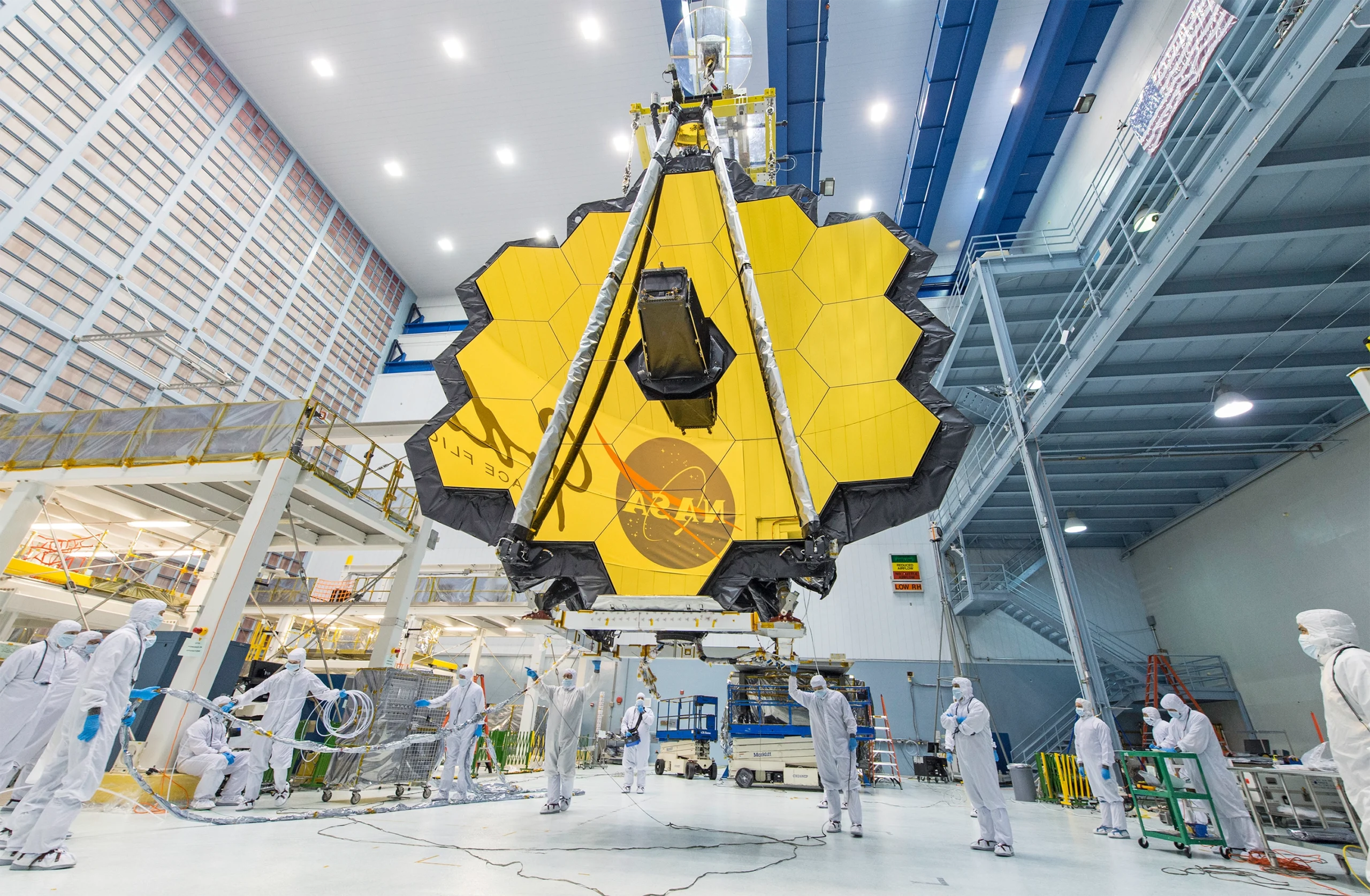 Photo of the giant, gold-plated mirror of the James Webb Space Telescope sitting in a clean room with people in bunny suits milling about.