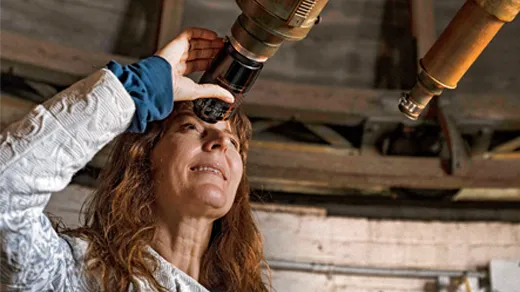 Lisa Kaltenegger, a woman with red hair, peers through the eyepiece of an antique telescope.