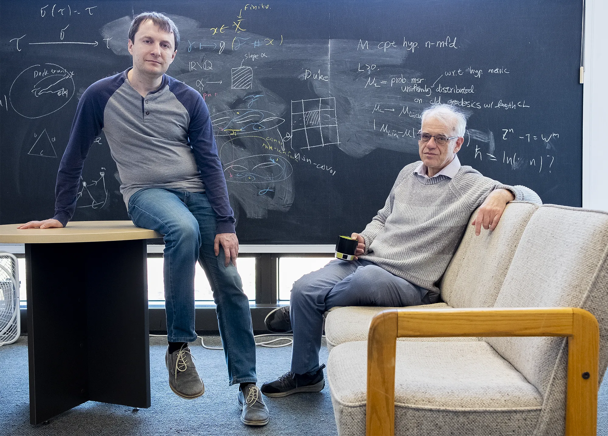 A photo of mathematicians Dima Dudko and Misha Lyubich sitting in front of a blackboard.