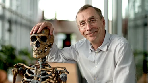 Photo of man with gold-rimmed glasses and a pin-striped button-up shirt posing with his hand on an early hominid skeleton