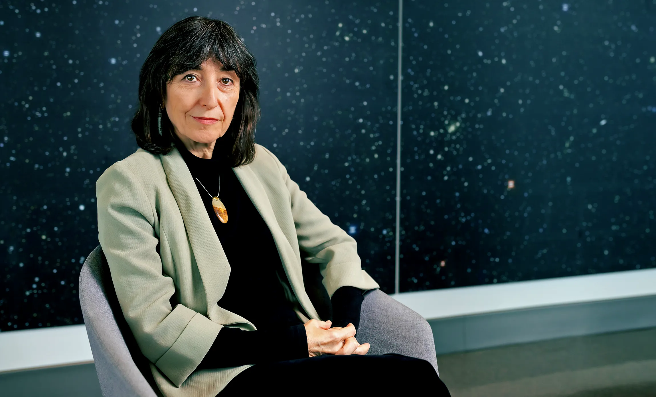 A woman with black hair is seated in an armchair in front of a wall-spanning photo of the night sky.